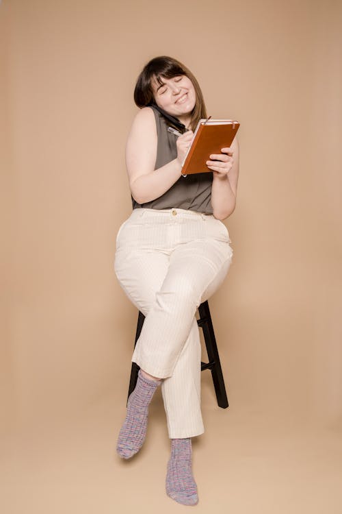 Full body of cheerful young female with closed eyes having phone call and taking notes in notebook against beige background