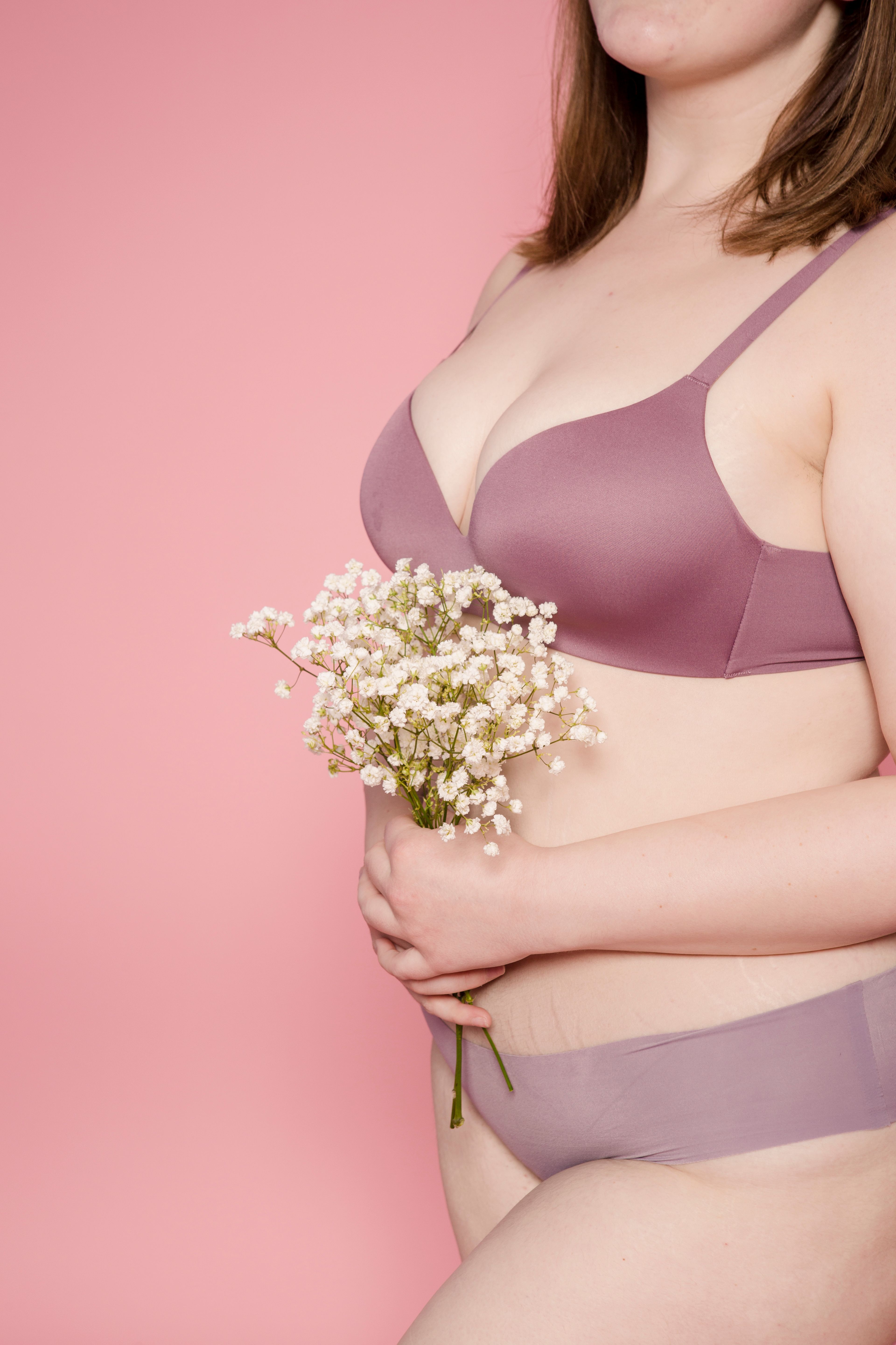 Side view of crop anonymous female in lingerie standing with bouquet of delicate flowers against pink background