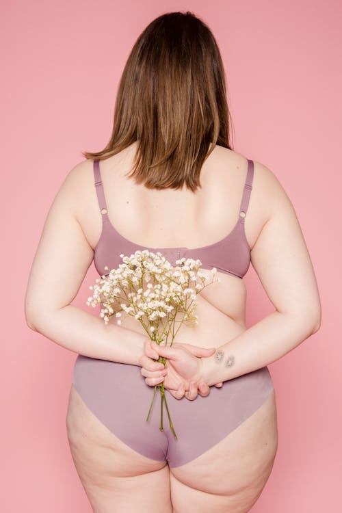 Back view of anonymous plump female in bra and panties demonstrating curves of body standing with bunch of fresh flowers against pink background