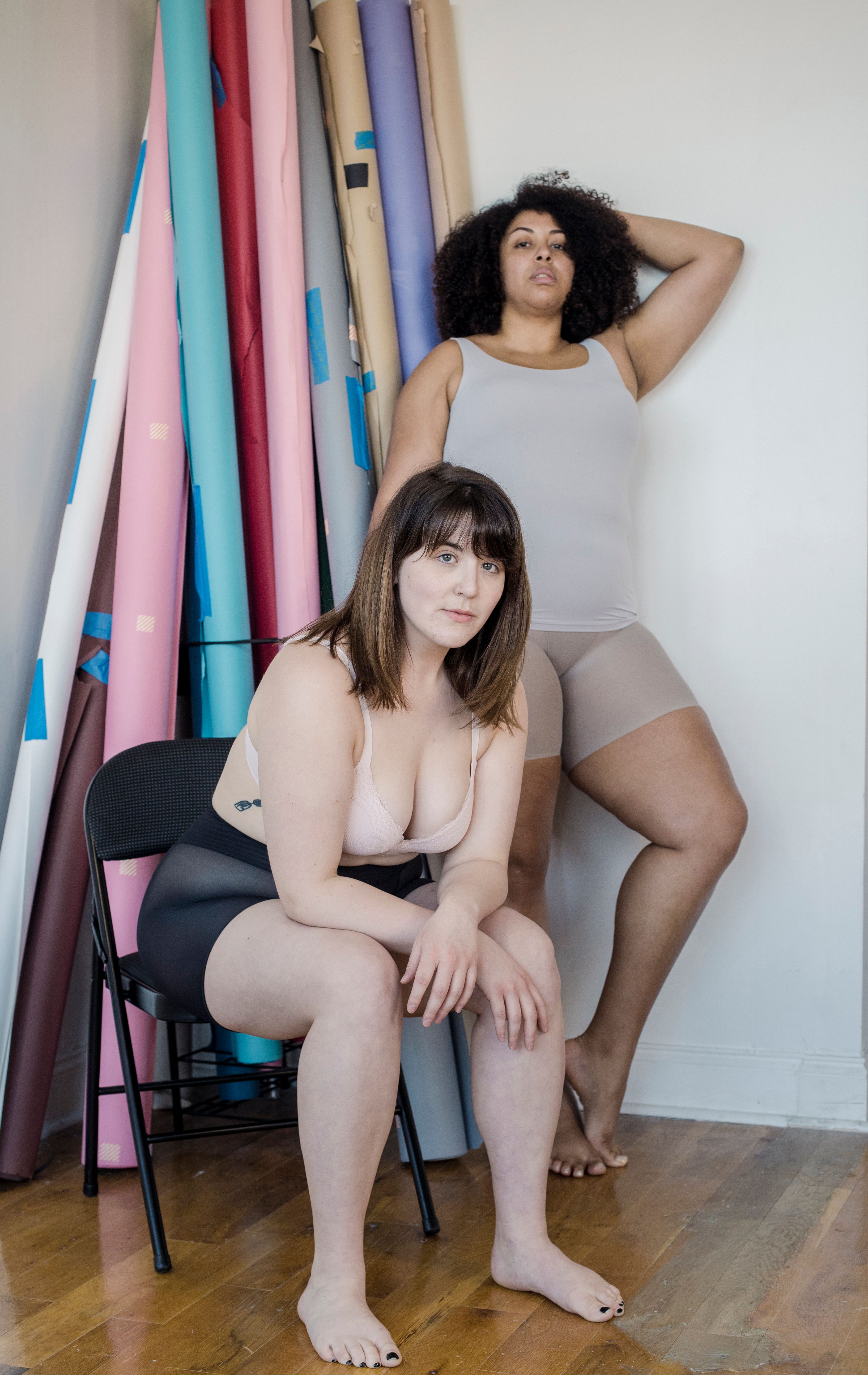 Full body of calm Asian woman and African American female in lingerie looking at camera while spending time in room with rolled wallpapers