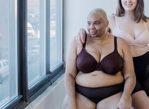 Crop plus size female and overweight African American woman in lingerie spending time together in light room near big window