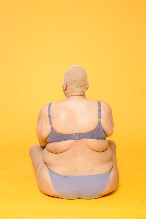 Free Back view of anonymous plump female model with short hair wearing lingerie sitting on floor on yellow background in studio Stock Photo
