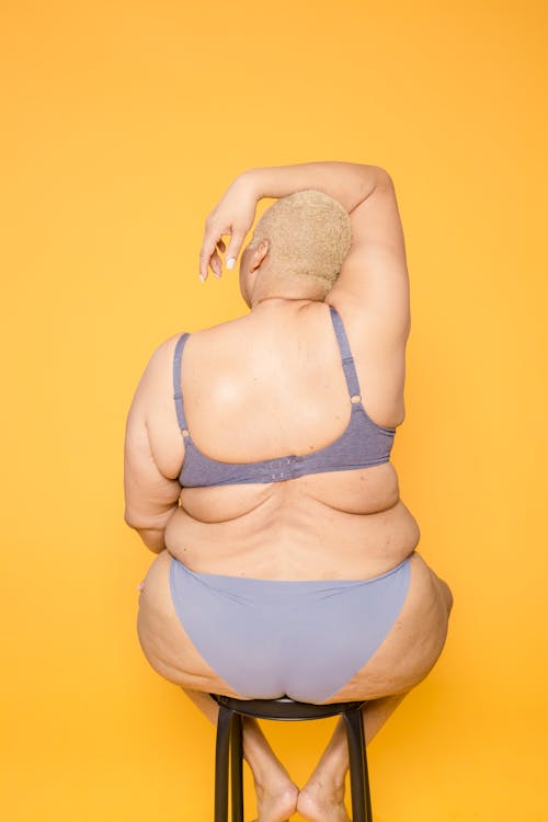 Back view of anonymous plus size female in lingerie sitting on chair with arm on head on yellow background in studio