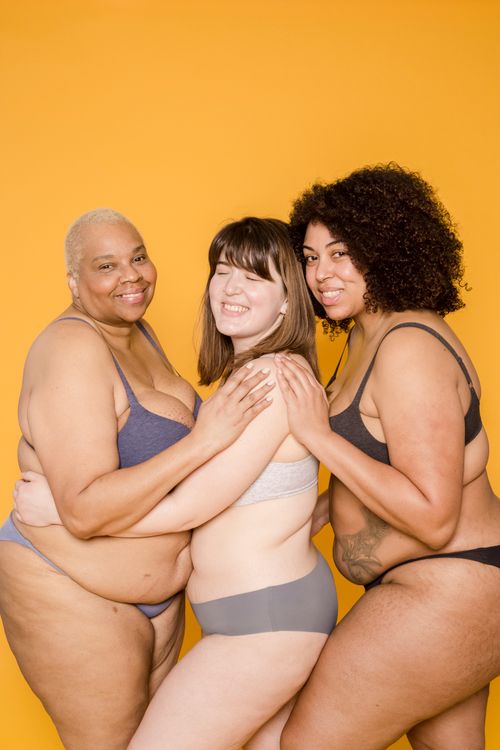 Free Happy multiracial plus size female models in lingerie embracing each other and looking at camera on yellow background in studio Stock Photo