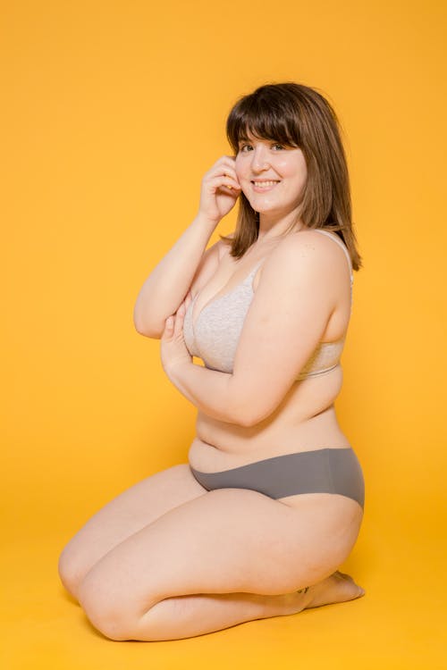 Free Full body of positive plump Asian female wearing lingerie looking at camera while sitting on yellow background in light studio Stock Photo