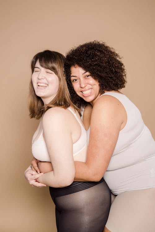 Free Side view of cheerful young overweight diverse female friends in underwear hugging and smiling happily against beige background Stock Photo