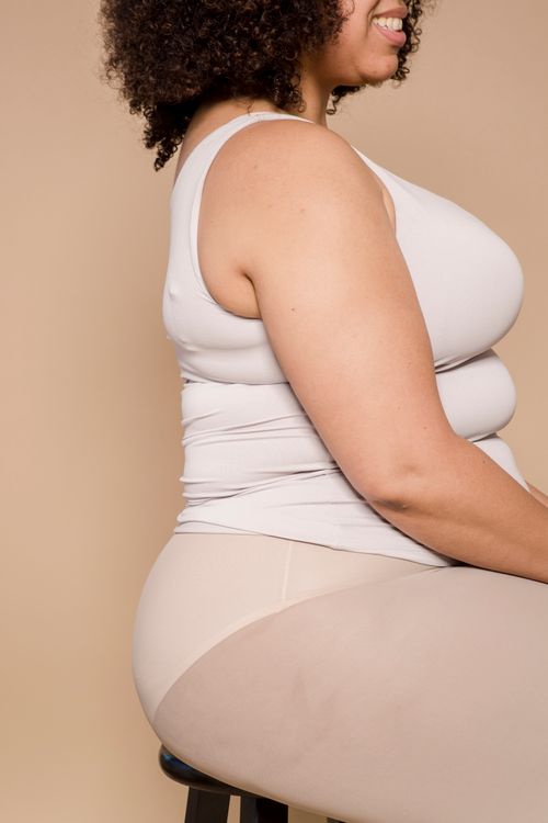 Free Side view of crop unrecognizable happy obese black lady with Afro hair in white top and leggings smiling while sitting on chair against beige background in studio Stock Photo