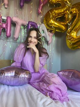 Woman in a purple dress sitting on a bed surrounded by colorful celebration balloons, smiling joyfully.