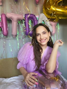 Smiling woman in a purple dress celebrates her birthday indoors with colorful balloons.