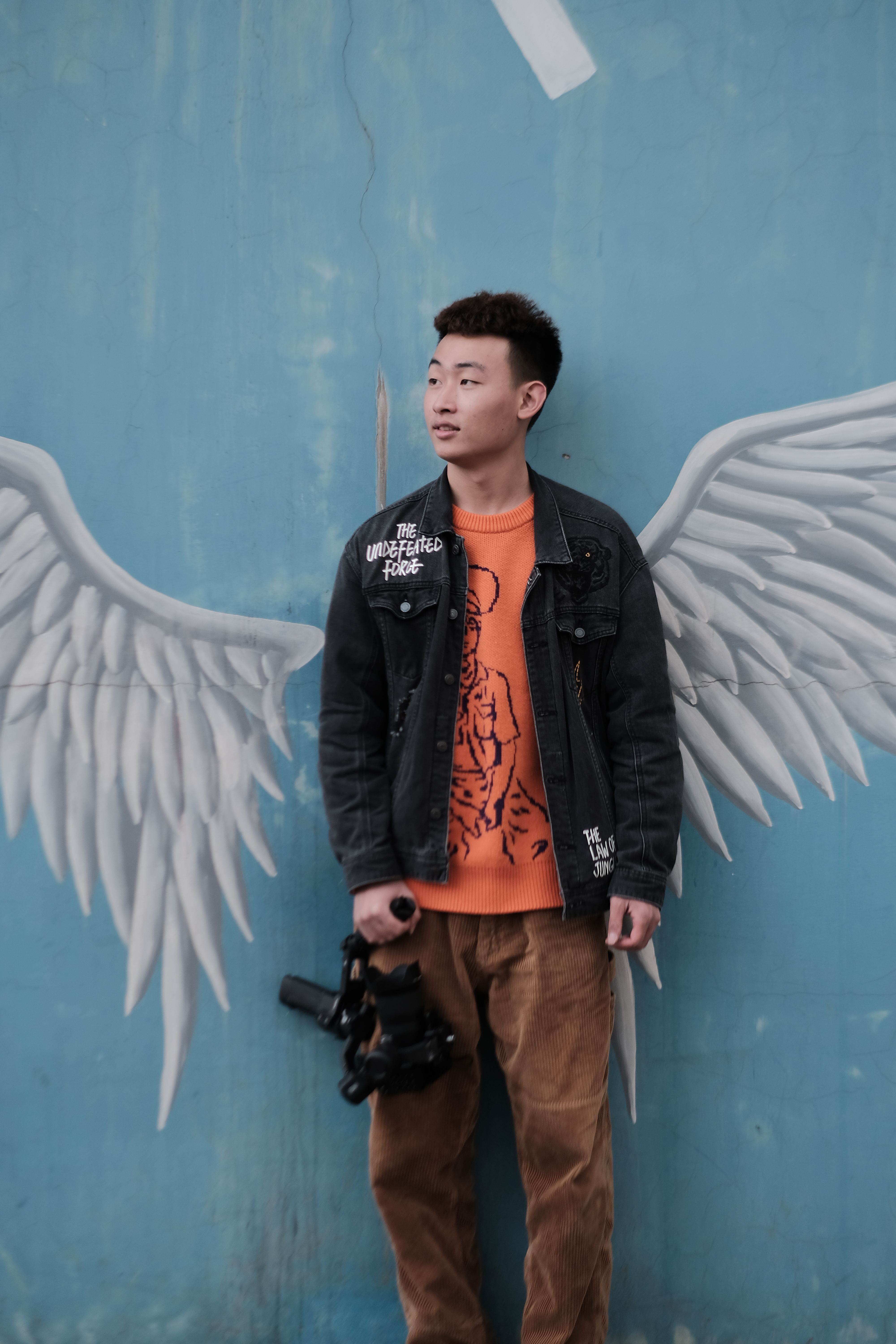 Young man with camera poses against a mural of angel wings on a blue wall.