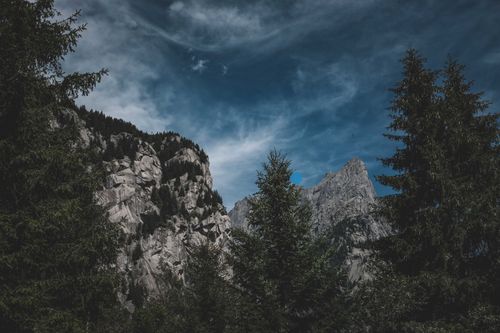 Free Majestic mountain scene with rugged cliffs and a dramatic blue sky, framed by dense pine trees. Stock Photo