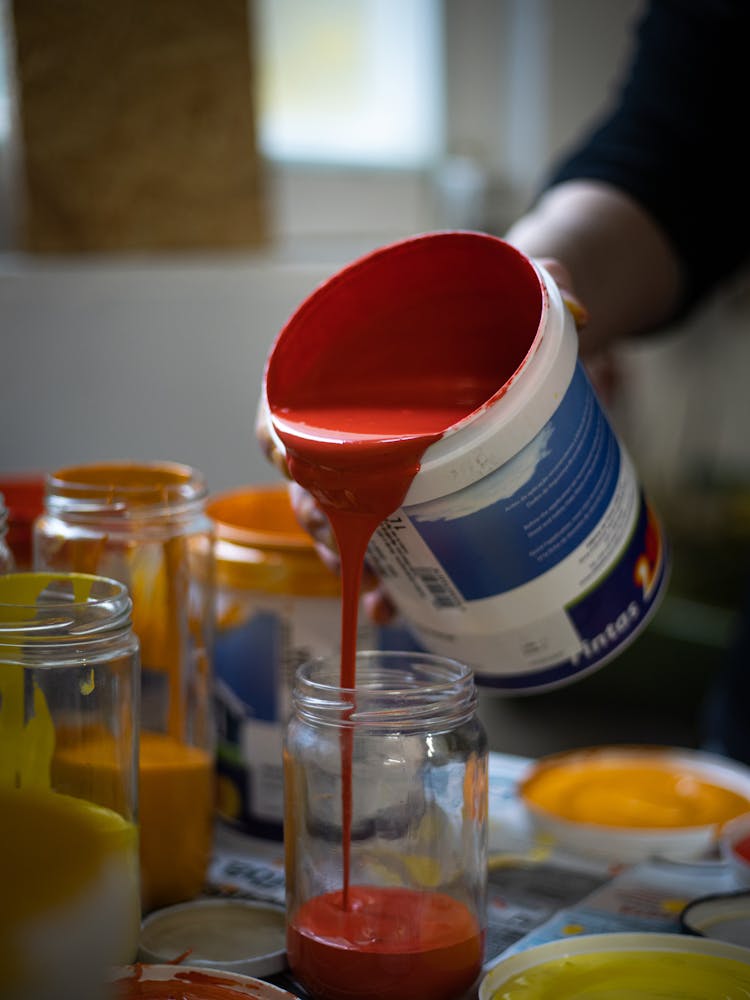 A Person Pouring Red Paint Into A Glass Jar