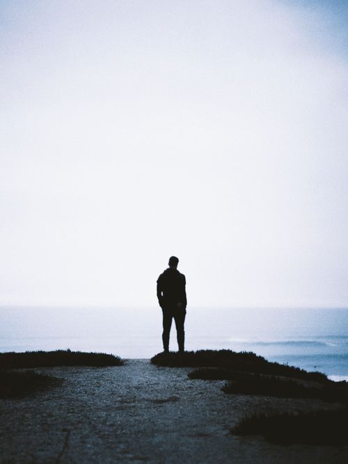 Free Silhouette of a person standing on a cliff above the ocean, surrounded by misty horizons. Stock Photo