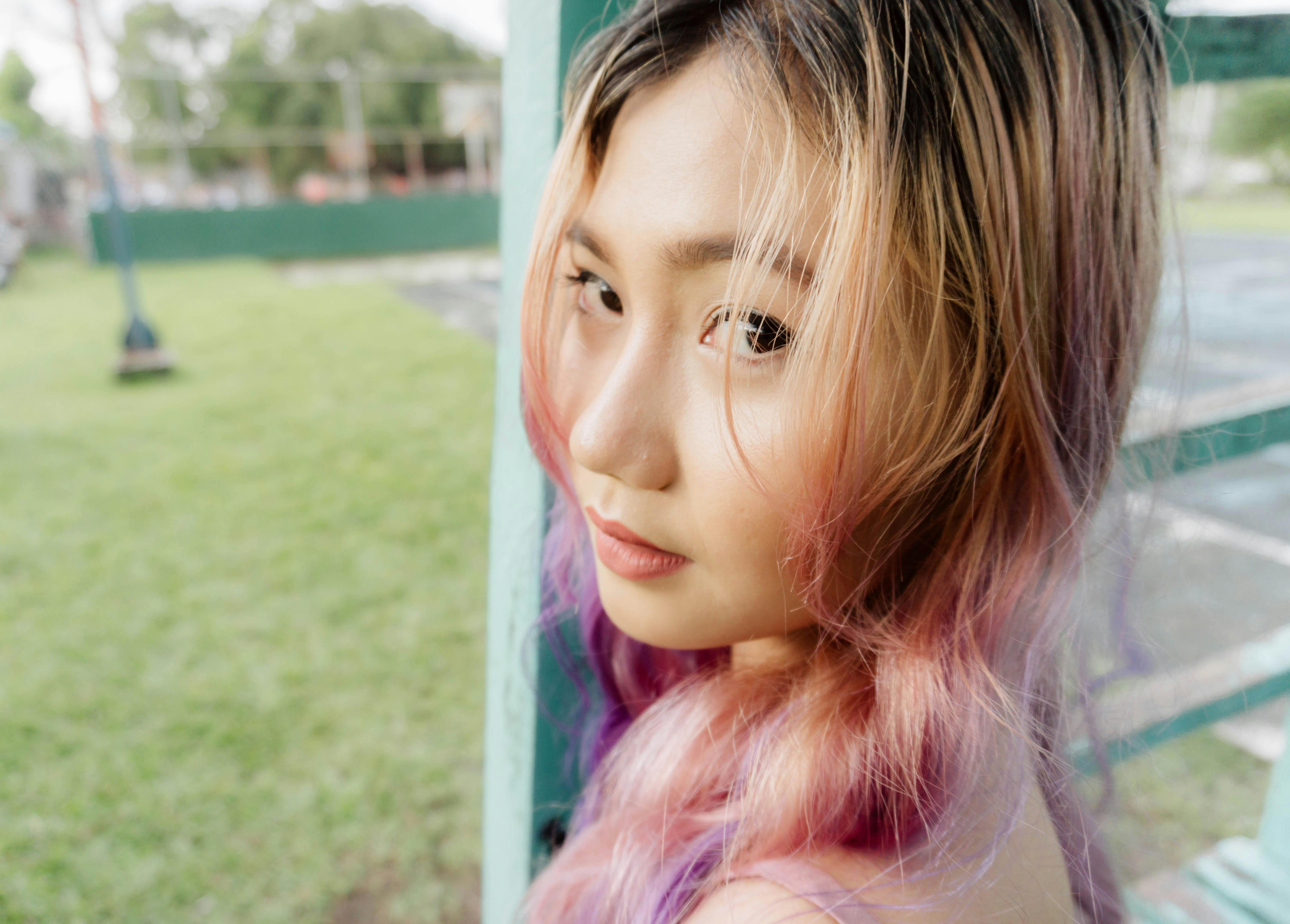 Asian girl with vibrant hair color leans against a green fence in a grassy outdoor setting.