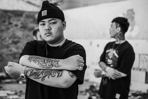 Black and white of young confident ethnic man showing tattoos while looking at camera against unrecognizable band in city