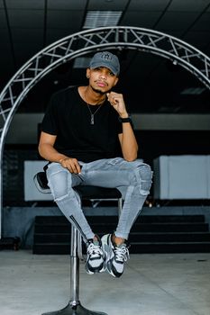 Young man in trendy attire sitting on a stool in an urban studio environment.