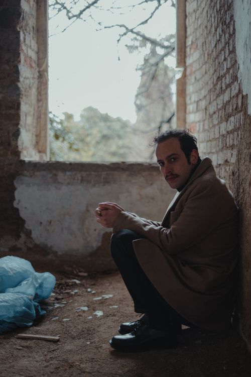 Free Side view full body of serious bankrupt male in stylish clothes looking at camera while sitting near wall in dirty ruined house Stock Photo