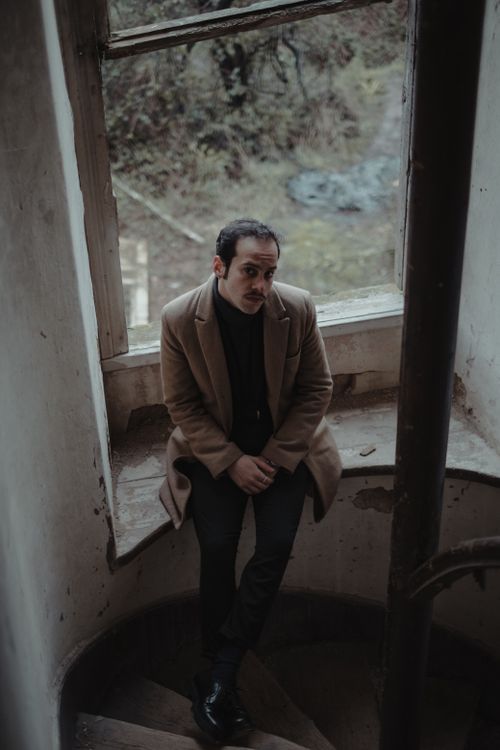 Free From above full body of serious male in elegant clothes looking at camera while sitting on dirty windowsill near staircase of weathered house Stock Photo