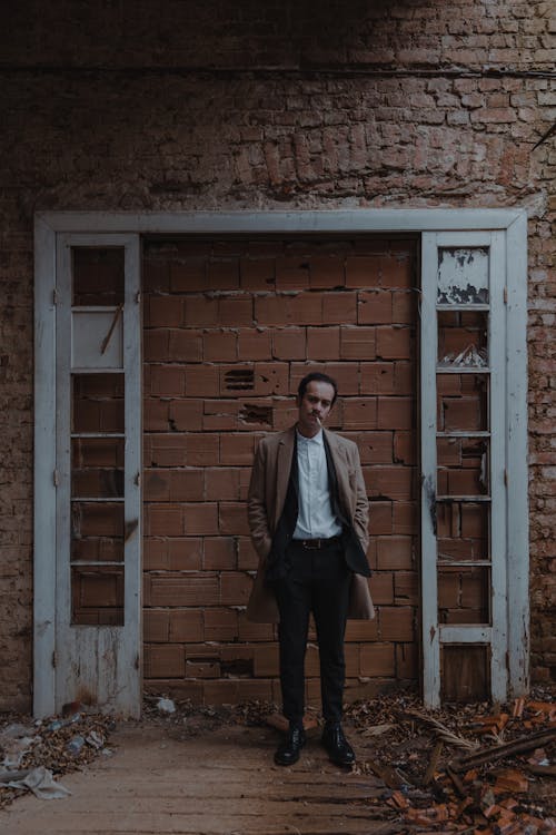 Full body of trendy male in elegant outfit smoking cigarette and looking at camera while standing near doorway blocked with brick wall
