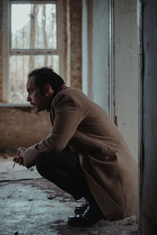 Free Side view full body of serious wistful male in stylish clothes squatting in weathered building with window and shabby walls Stock Photo