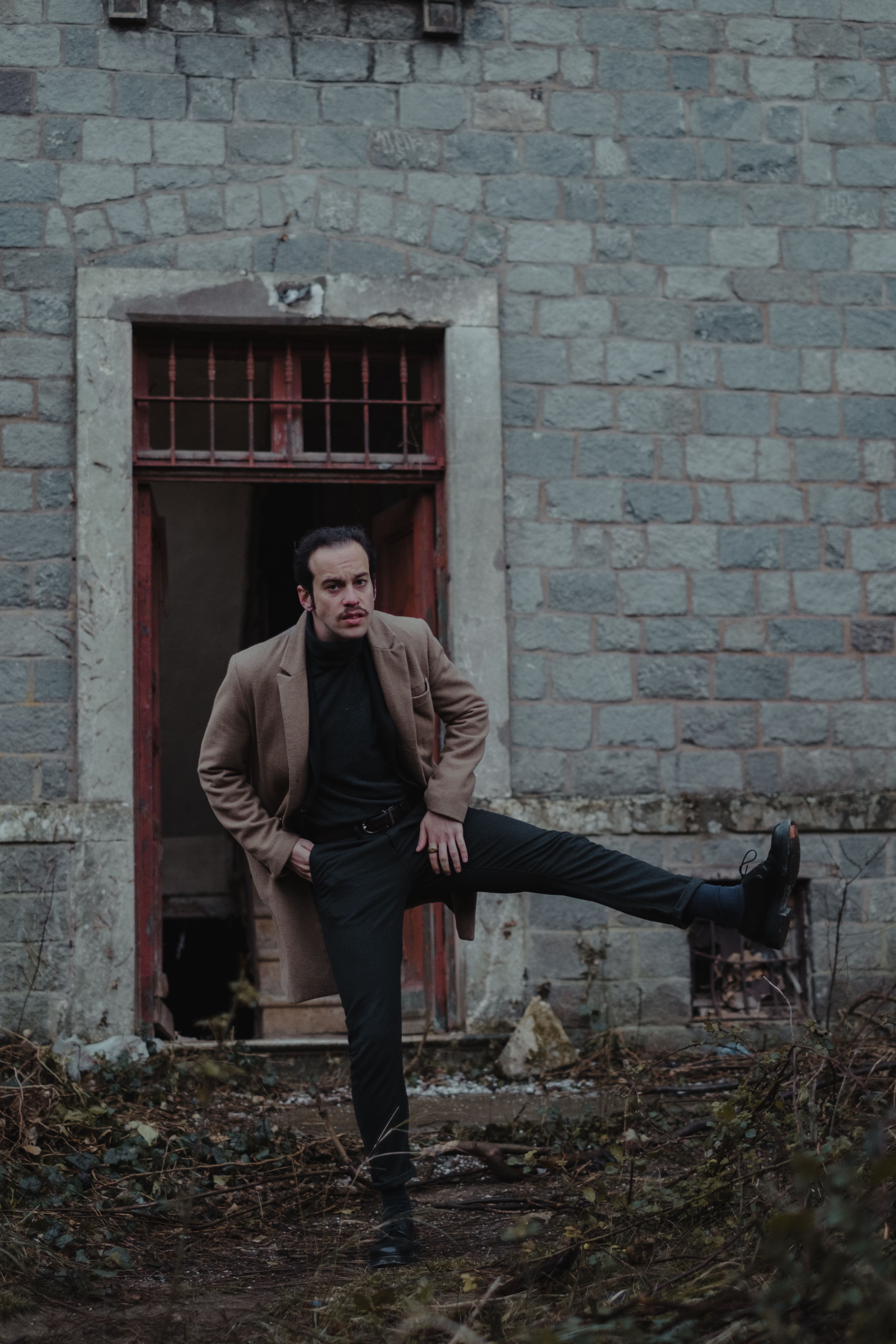 Full body of male in elegant outfit with raised leg looking at camera while standing near shabby building wall stone wall and doorway