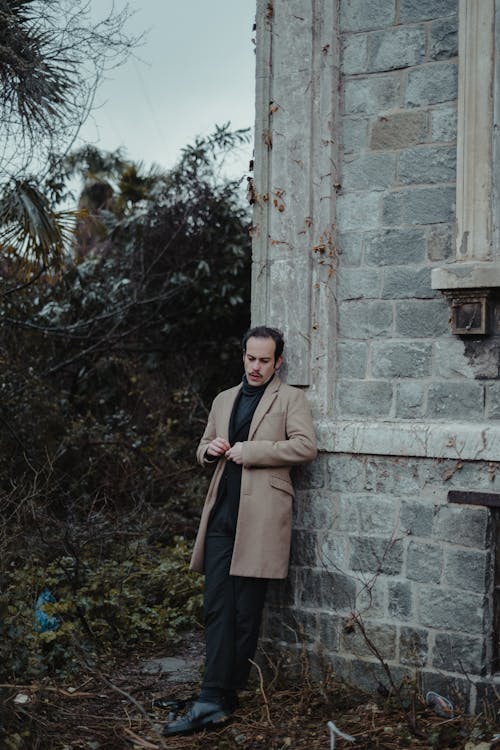 Full body of stylish male in elegant coat leaning on shabby concrete building and looking down