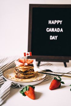 Stack of pancakes with maple syrup and strawberries, celebrating Canada Day with festive flag.