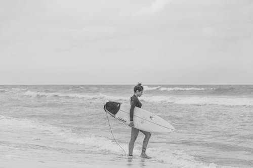 Grayscale Photo of Woman Holding Surfboard on Beach