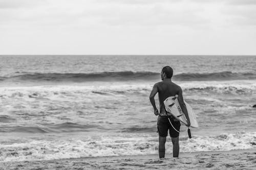 Grayscale Photo of Man Holding Surfboard on Beach