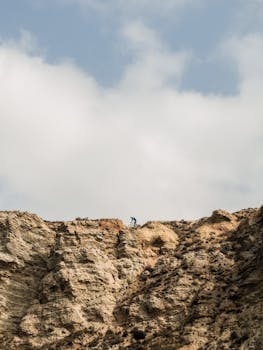 A lone mountain biker navigates the rocky cliffs with white clouds above.