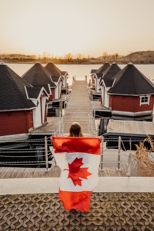 Back View Shot of a Woman Holding a Canadian Flag while Standing on a Wooden Dock Near Villas