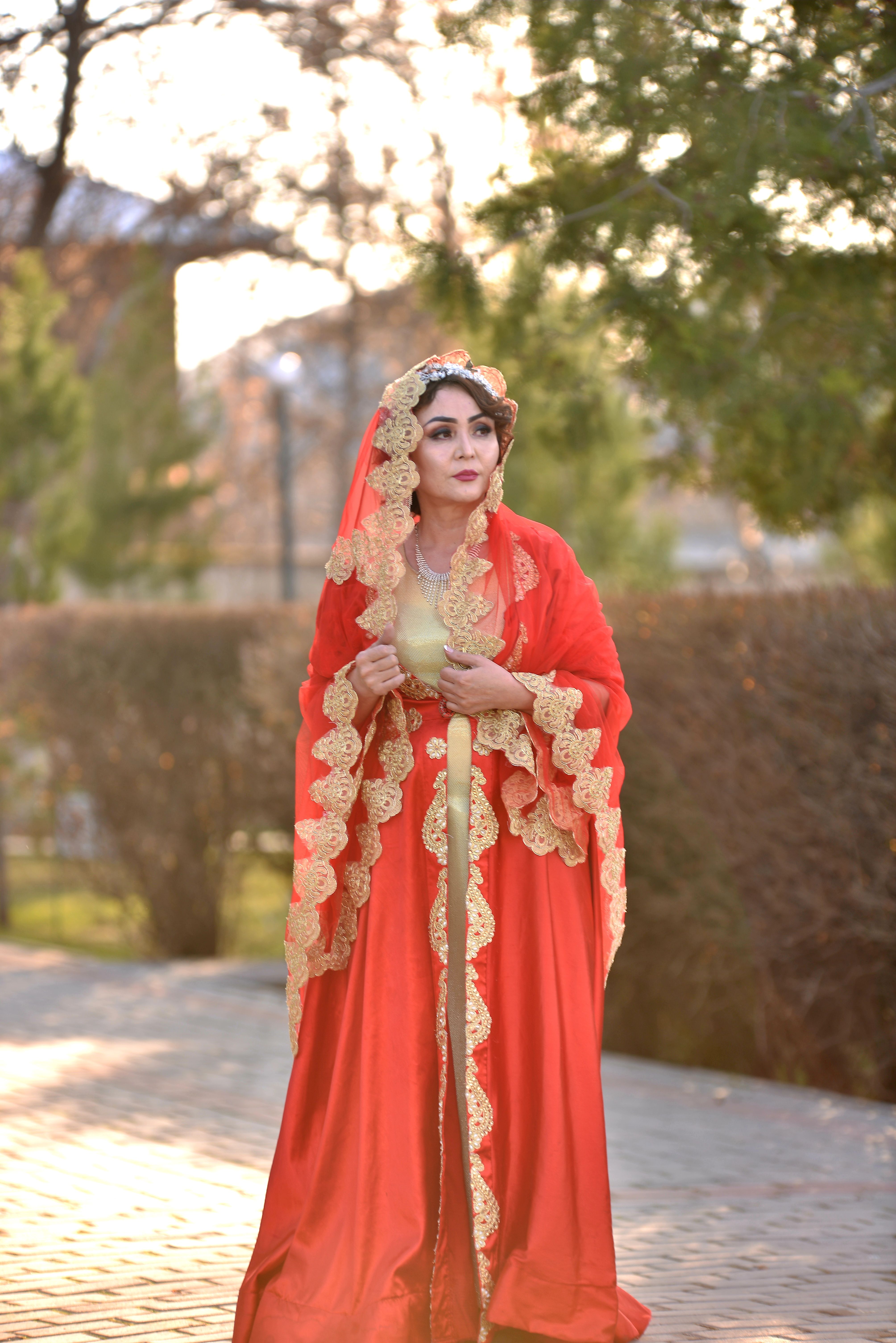 Woman in an elaborate red and gold traditional dress standing outside, exuding elegance.