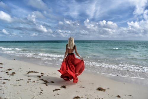 女人穿著紅色的裙子，走在海邊，在藍色和白色的天空下