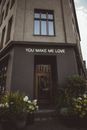 House exterior with You Make Me Love title above entrance door against blossoming flowers in city