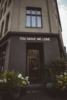 House exterior with You Make Me Love title above entrance door against blossoming flowers in city