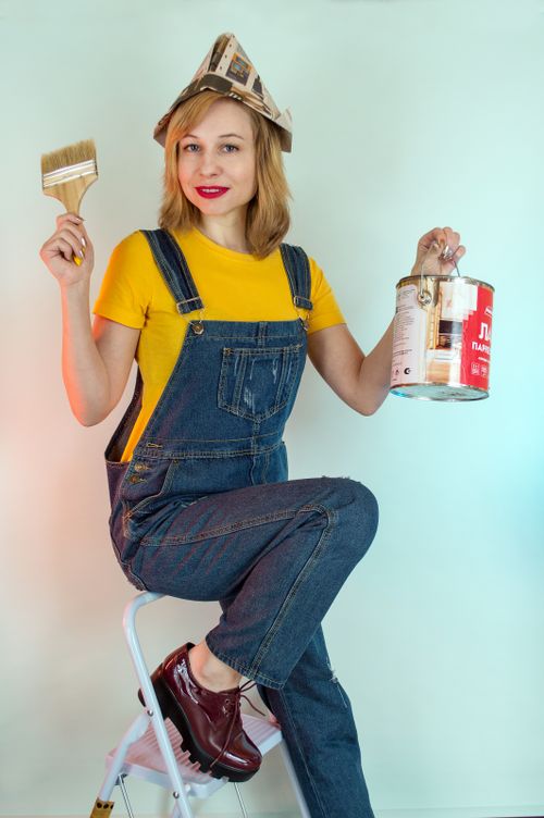 Free Cheerful female painter in denim workwear and paper hat showing brush and paint can while sitting on ladder and looking at camera Stock Photo