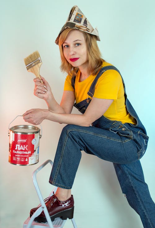 Content female painter wearing denim jumpsuit and paper hat leaning on ladder and holding paint can and brush while looking at camera