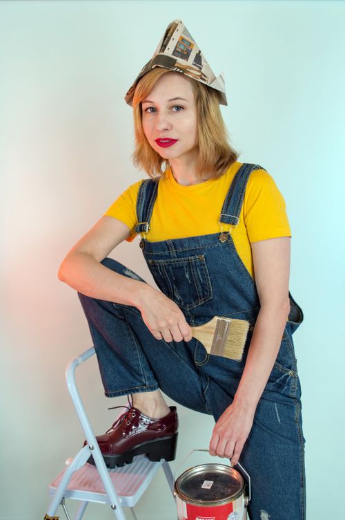 Free Glad female decorator wearing denim workwear and paper hat holding brush and paint can and looking at camera while leaning on ladder against white wall Stock Photo