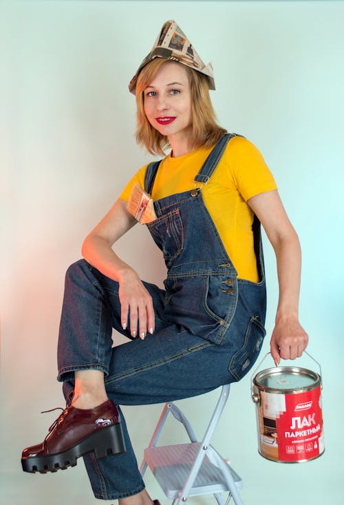 Happy female in denim workwear and paper hat holding pain can and sitting on ladder against white wall while looking at camera contentedly