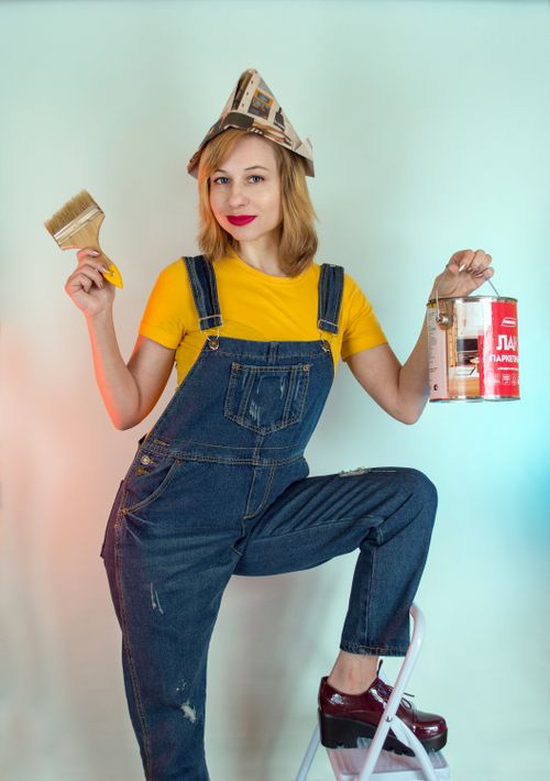 Free Glad female painter wearing denim jumpsuit and paper hat demonstrating brush and paint can while leaning on ladder and looking at camera Stock Photo