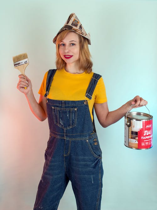 Content young female in denim workwear and paper hat holding brush and paint can while standing against white wall and looking at camera with smile