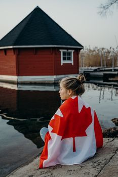 A contemplative young woman sits by the water wrapped in a Canadian flag in Toronto.