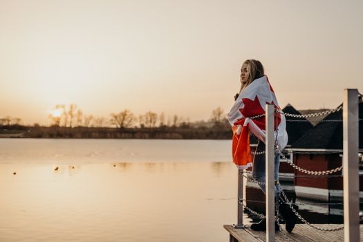 A woman wrapped in a Canadian flag stands on a dock at sunset, gazing at the serene lake in Toronto.