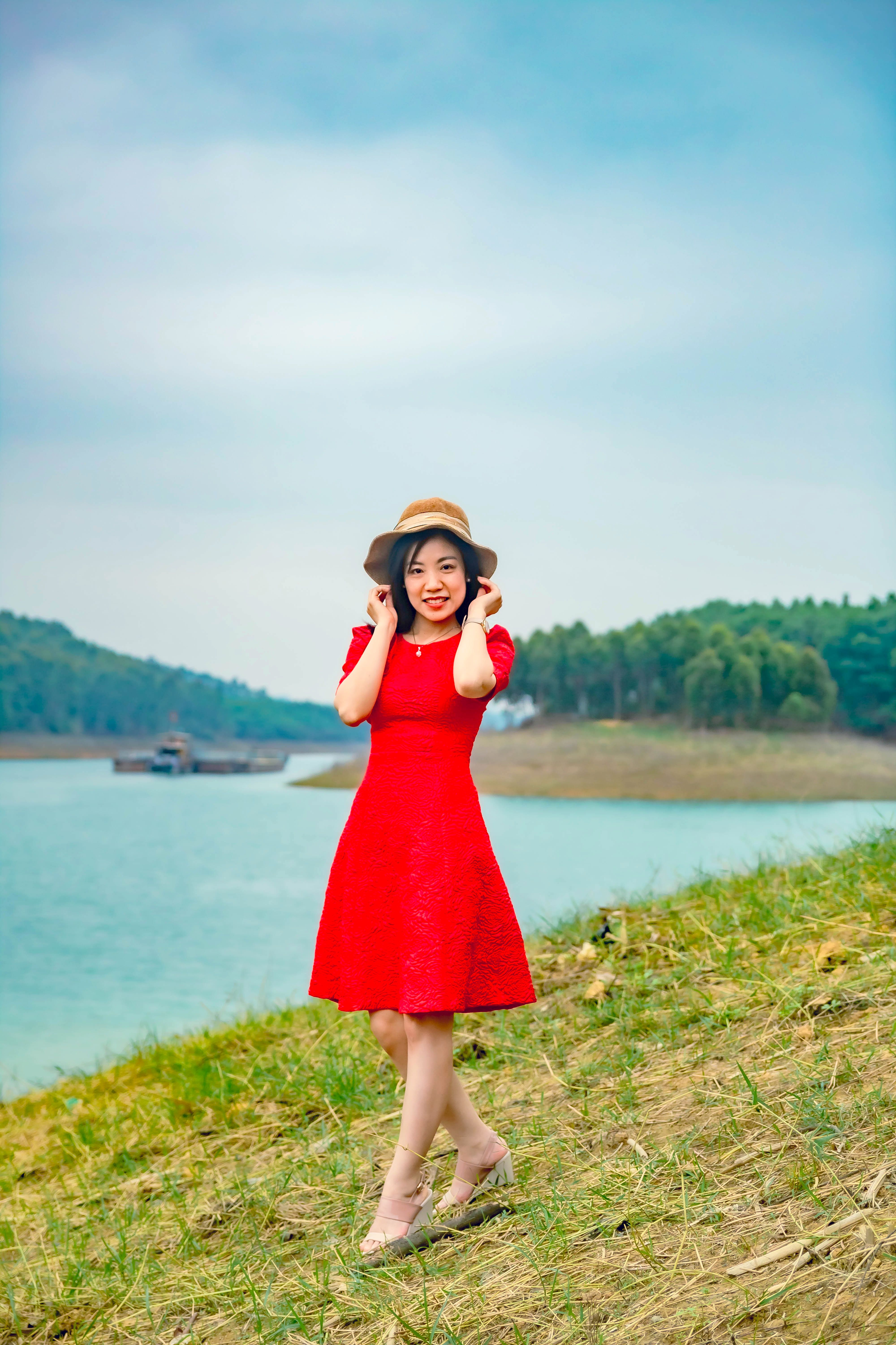 Smiling woman in a red dress, brown hat, standing by a picturesque lake.