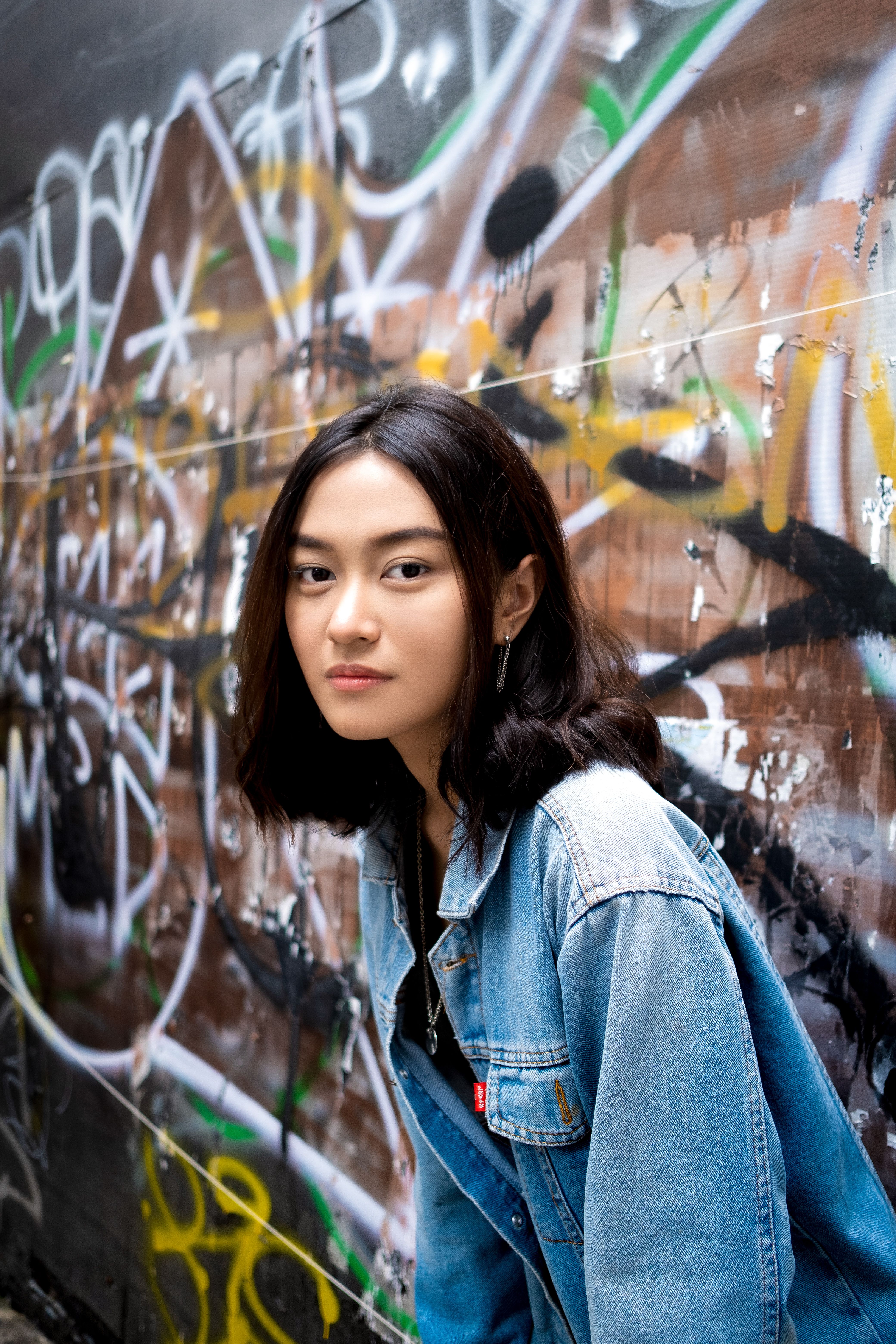 Asian woman in denim jacket poses by vibrant graffiti wall, showcasing street style fashion.