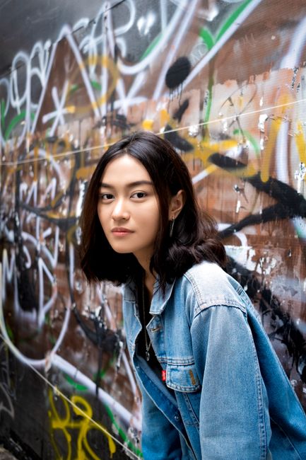 Asian woman in denim jacket poses by vibrant graffiti wall, showcasing street style fashion.