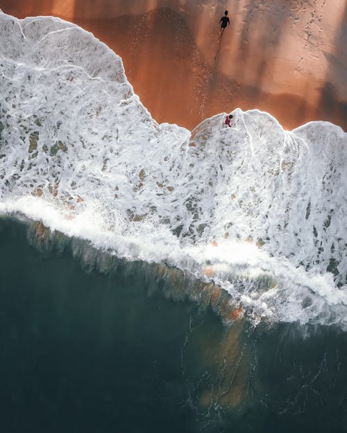 Drone view of travelers on sandy beach of powerful blue ocean with foamy waves in tropics