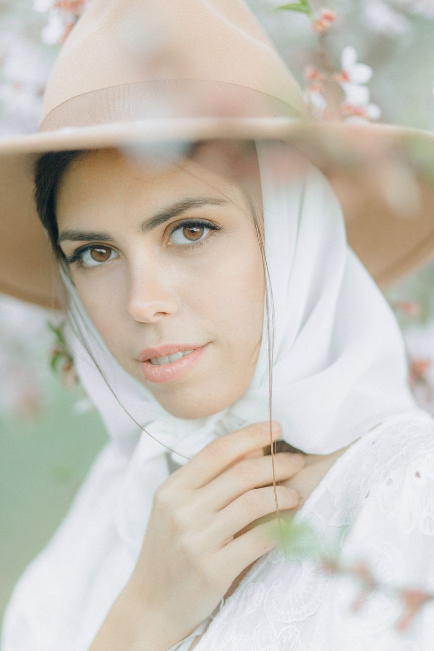 Portrait of a woman wearing a white headscarf and brown fedora among cherry blossoms.
