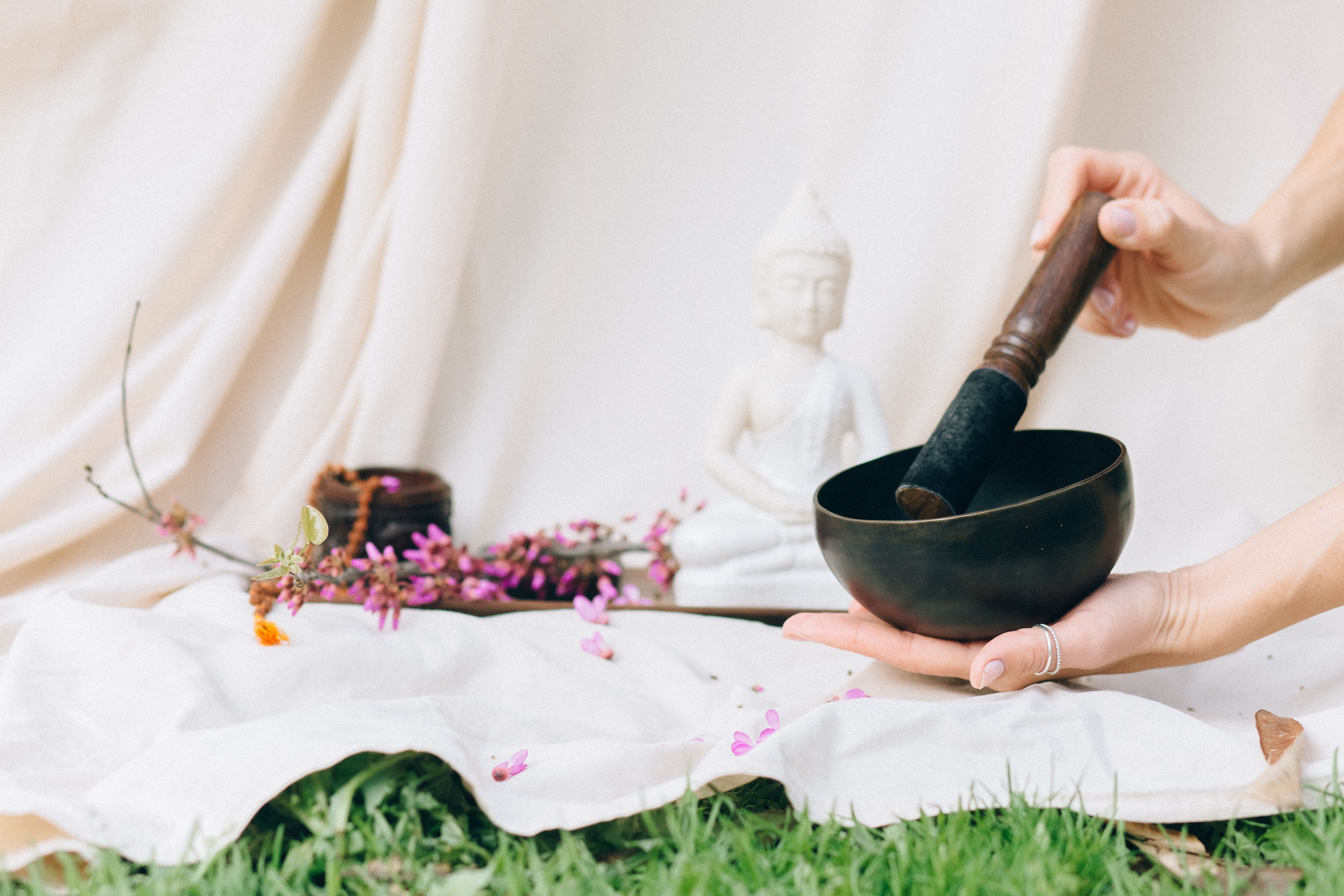Hands holding a Tibetan singing bowl for meditation in a serene setting with a Buddha figurine and flowers.