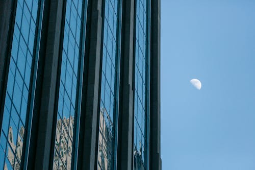 From below of exterior of modern high rise building with glass mirrored walls against cloudless blue sky with moon in daytime